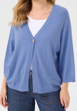 Femme portant un cardigan bleu clair ample avec un bouton fermé sur un haut blanc et un pantalon en jean bleu foncé.