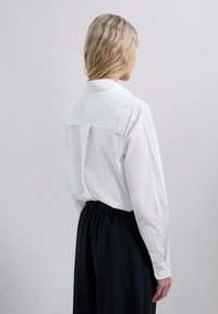 Weißes Hemd mit Rückenfalten, langen Ärmeln und Button-Ärmeln, kombiniert mit dunkel gestreiften Hosen, von hinten betrachtet.