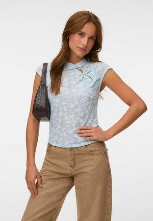 Femme portant un haut en dentelle florale bleu clair avec des boutons noués, un pantalon beige et un sac à épaule texturé foncé, posant avec la main sur la hanche.