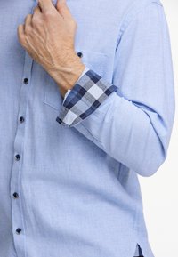 Ljusblå skjorta med knappar och vänster bröstficka, med ett mönster i marinblått och ljusblått rutmönster på muddarna. Tillverkad av bomull, med en mjuk textur.