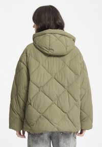 Chaqueta de plumón acolchada de color verde oliva con capucha, que presenta un patrón de diamantes y un cordón elástico en el dobladillo. Vista posterior solamente.