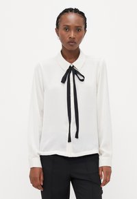 Blouse blanche avec un col et un nœud en ruban noir. Dotée de manches longues, d'une fermeture à boutons et d'une texture de tissu lisse et légère.