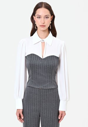 Femme portant un chemisier blanc à manches longues avec un corset gris à rayures fines et un pantalon assorti sur un fond clair uni.