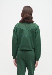 Άτομο με καστανά μακριά μαλλιά, ντυμένο με πράσινο μακρυμάνικο μπλουζάκι και ταιριαστά πράσινα παντελόνια, στραμμένο μακριά σε λευκό φόντο.