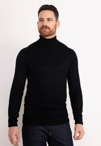 Maglione a collo alto nero realizzato in un materiale liscio ed elastico; design aderente con maniche lunghe e polsini a costine. Indossato con jeans blu.