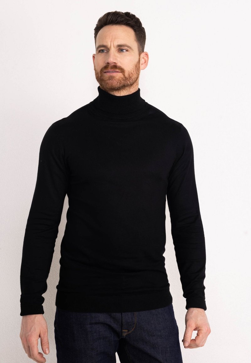 Maglione a collo alto nero realizzato in un materiale liscio ed elastico; design aderente con maniche lunghe e polsini a costine. Indossato con jeans blu.