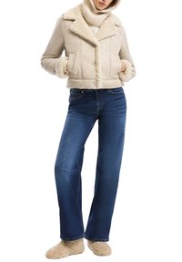 Vrouw in een beige schapenvachtjack, crèmekleurige coltrui, blauwe jeans en beige fluffy slippers, staande met handen in haar zakken.