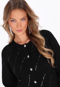 Cardigan nero in maglia con un motivo testurizzato, decorazioni in paillettes argentate e chiusure con bottoni rotondi. Il tessuto testurizzato aggiunge profondità.