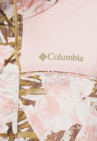 Κοντινό πλάνο ροζ και καφέ μπουφάν με λουλουδάτο σχέδιο της Columbia που δείχνει το λογότυπο της μάρκας και ένα μπεζ κορδόνι φερμουάρ.