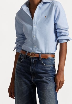 Personne portant une chemise bleu clair à boutons avec les manches retroussées, rentrée dans un jean bleu foncé, et une ceinture en cuir marron avec une boucle dorée.