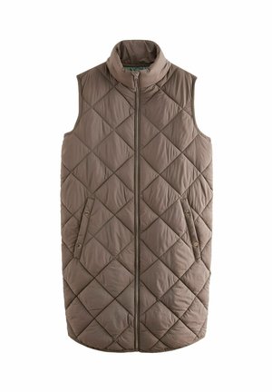 Femme portant un long gilet matelassé gris sans manches sur une chemise sombre à manches longues, les mains dans les poches du gilet, debout devant un fond uni.