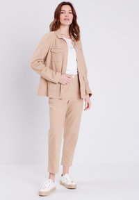 Beige linnen jas met een kraag en knoopsluiting, gecombineerd met bijpassende beige broek. Witte kanten top en witte sneakers met een gevlochten zool.