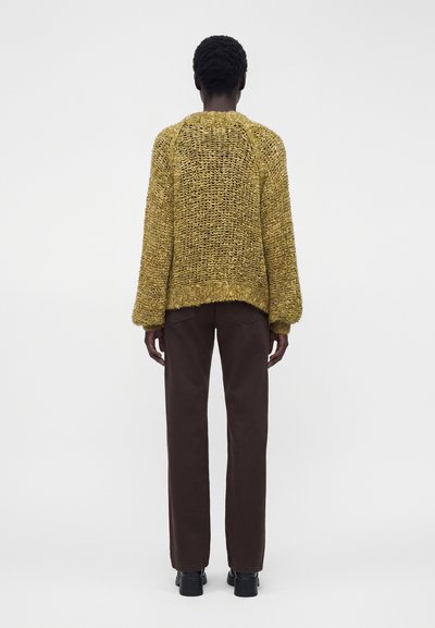 Personne portant un pull jaune moutarde en maille lâche, un pantalon droit marron foncé et des chaussures noires, debout de dos sur un fond uni.
