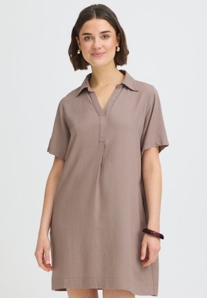 Fiatal nő rövid barna hajjal, bézs rövid ujjú, galléros ruhában és sötét karkötővel, mosolyogva, egyszínű háttér előtt.