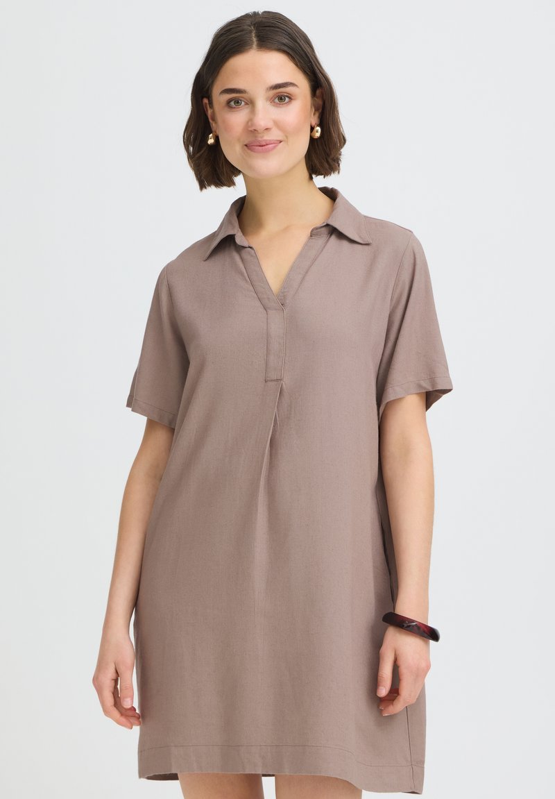 Jeune femme aux cheveux bruns courts portant une robe beige à manches courtes avec col et un bracelet sombre, souriante devant un fond uni.