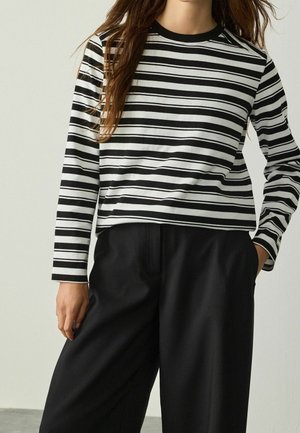 Femme portant un haut rayé horizontal noir et blanc à manches longues et un pantalon noir ample, debout avec une main dans la poche.