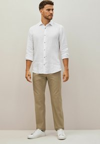Vit linneskjorta med knappar och uppvikta ärmar, kombinerad med ljusbeige byxor och vita sneakers. Slät textur, avslappnad design.