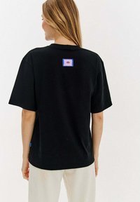 Czarna koszulka z krótkim rękawem z prostokątną niebiesko-różową łatą z logo oka na plecach, wykonana z miękkiego materiału bawełnianego.