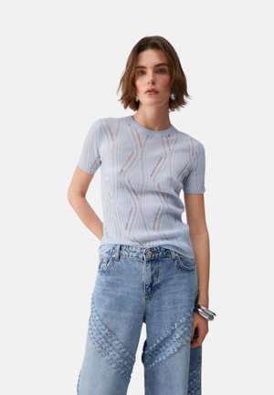 Giovane donna con capelli castani corti che indossa un maglione a maniche corte azzurro chiaro con motivi e jeans blu a vita alta e a gamba larga.