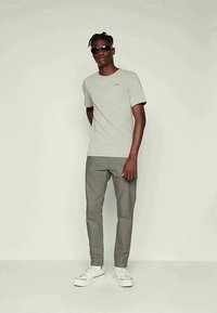 Camiseta cinza claro com mangas curtas, calças cinzas e ténis brancos. Modelo com óculos de sol está encostado a um fundo neutro.