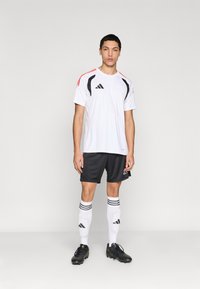 Λευκό αθλητικό μπλουζάκι με μαύρες και κόκκινες προστευτικές λεπτομέρειες, μαύρο σορτς και λευκές κάλτσες με μαύρες ρίγες, φορεμένα με μαύρα ποδοσφαιρικά παπούτσια.