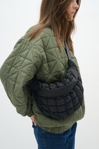 Veste matelassée vert olive assortie à un sac à main noir bouffant de forme arrondie et à la texture lisse, maintenu par une sangle.