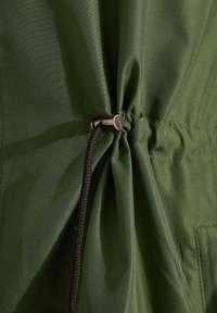Dettaglio di una giacca in tessuto verde con una sezione arricciata fissata da un cordino nero e un fermaglio in metallo argento.