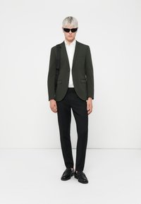 Mørkegrøn blazer over en hvid skjorte, sorte bukser og sorte sko. Modellen bærer sorte solbriller med platinblond hår.