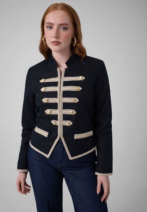 Mujer con cabello rojo que lleva una chaqueta de estilo militar negra con adornos y botones trenzados dorados, combinada con vaqueros azul oscuro, de pie frente a un fondo gris.