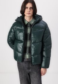 Chaqueta de abrigo verde con una textura brillante, equipada con capucha y cierre de cremallera, llevada sobre una camisa de cuadros. Combinada con jeans oscuros.