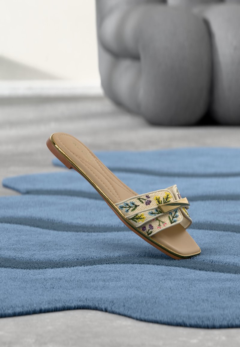 Sandale plate beige avec broderie florale et boucle dorée sur un tapis bleu texturé, avec un canapé gris flou en arrière-plan.