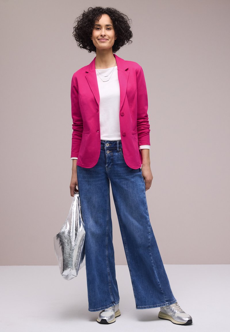 Femme aux cheveux bouclés portant un blazer rose, une chemise blanche, un jean large bleu, des baskets argentées et tenant un sac fourre-tout argenté métallique.