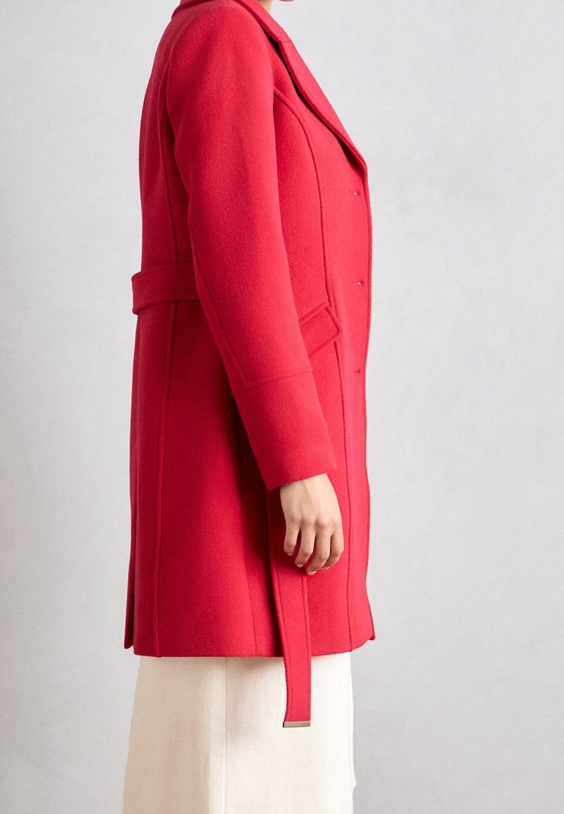 Personne portant un manteau en laine rouge cintré avec des détails de ceinture sur une jupe blanche, vue de profil sur un fond gris uni.