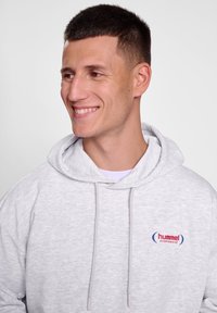 Hoodie gris avec cordon de serrage, arborant un petit logo "hummel sportswear" rouge et bleu sur la poitrine gauche. Texture douce et coupe décontractée.