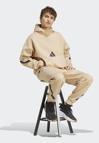 Felpa beige con cappuccio e pantaloni della tuta con accenti neri e logo a tre strisce, realizzati in materiale morbido. Scarpe sportive nere con design testurizzato.