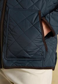 Prešívaná tmavomodrá bunda s hnedými kordurovými detailmi, bočnými vreckami a zipsovým uzáverom. Hladká textúra s kovovým vzorom.