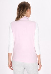 Gilet senza maniche di colore rosa chiaro con colletto alto, realizzato in morbido materiale in fleece. Indossato sopra una camicia bianca a maniche lunghe, presenta una texture liscia.
