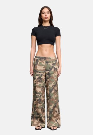 Mujer de pie recta, vistiendo una camiseta negra corta, pantalones anchos de camuflaje y sandalias negras sobre un fondo claro y liso.