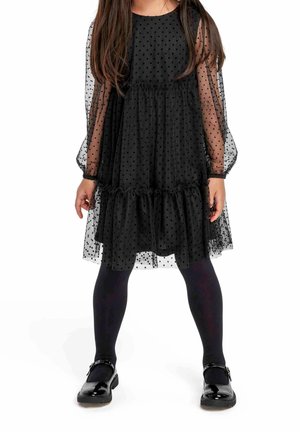 Robe noire à pois avec manches longues en tulle transparent, volants en étages à l'ourlet, associée à des collants noirs et des chaussures vernies à boucle.