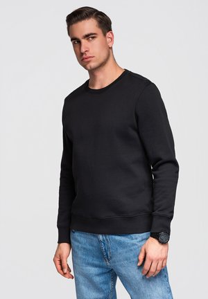 Jeune homme portant un sweat-shirt noir et un jean bleu, debout devant un fond clair uni, regardant sur le côté.