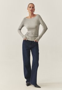 Graues Langarmshirt mit weitem Ausschnitt, kombiniert mit dunkelblauen weit geschnittenen Jeans und schwarzen spitzen Schuhen. Glattes Material, figurbetont und lässiger Stil.