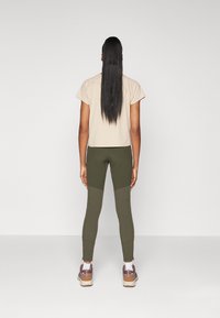 Beige t-shirt med korta ärmar, kombinerad med olivgröna leggings som har texturerade paneler. Modellen bär sportskor i en dämpad färgpalett.