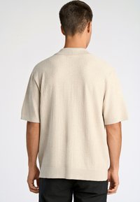 Korte mouwen, beige polo met een ribbelstructuur, voorzien van een platte kraag en een ontspannen pasvorm, achterkant weergegeven tegen een effen achtergrond.