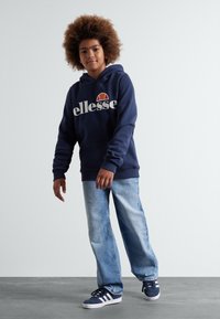 Tamno plava dukserica s kapuljačom s "ellesse" logotipom, u kombinaciji s svijetloplavim širokim trapericama i tamnoplavim tenisicama s bijelim prugama. Tekstura kose, neutralna pozadina.