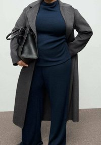 Personne portant une tenue bleu foncé et un long manteau gris, tenant un sac à main en cuir noir avec une main sur la hanche, debout devant un fond uni.