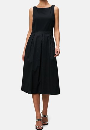 Vrouw draagt een mouwloze zwarte midi-jurk met een getailleerde taille en geplooide rok, gecombineerd met zwart-witte sandalen met open teen.