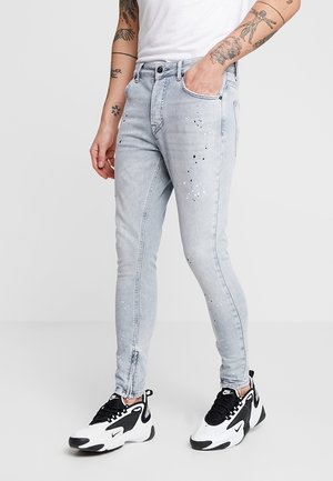 Schmal geschnittene hellgraue Jeans mit schwarzen und weißen Farbspritzern, getragen mit schwarzen und weißen Nike-Sneakern und einem weißen Hemd, das tätowierte Arme zeigt.