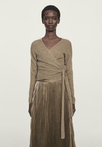 Beige Woll-Wickelsweater mit langen Ärmeln, kombiniert mit einem metallisch gefalteten goldenen Rock. Glatte Textur und gebundenes Detail in der Taille.