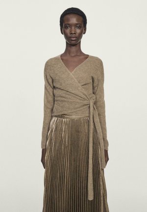 Beige Woll-Wickelsweater mit langen Ärmeln, kombiniert mit einem metallisch gefalteten goldenen Rock. Glatte Textur und gebundenes Detail in der Taille.