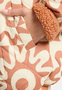 Hand met gepolijste nagels in de zak van een gewatteerde jas met abstracte zonpatronen en een fuzzy manchette in warme beige tinten.